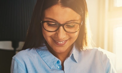Young smiling woman in light blue shirt wearing glasses, sat in a sunlit lounge, looking at laptop