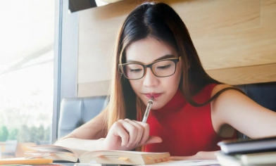 Woman sat in a well-lit café, holding pen to her mouth while reading from a book for her studies